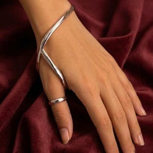 May include: A polished silver-tone hand bracelet featuring a curved, fluid design that wraps around the wrist and connects to a simple, smooth band worn around the thumb. The metal has a reflective, high-shine finish throughout the piece.