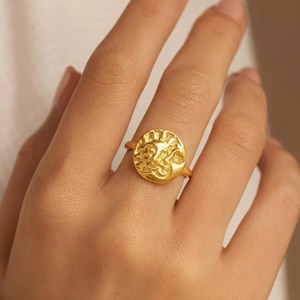 Può includere: Anello dorato con un design circolare strutturato con un motivo a raggiera. L'anello è indossato su un dito, mostrando i suoi dettagli intricati e la calda tonalità metallica. Il gambo dell'anello è sottile e delicato.