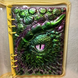 May include: A ring-bound journal featuring a three-dimensional, iridescent green and purple dragon eye cover with textured scales and a prominent horn. The journal is encased in a clear plastic protective sleeve with yellow trim and a yellow zipper.