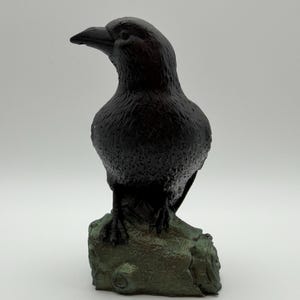 May include: A black crow figurine with a textured, matte finish perched on a weathered, green-toned log base. The bird is depicted in a standing position with its head turned slightly to the left, showcasing detailed feather patterns.