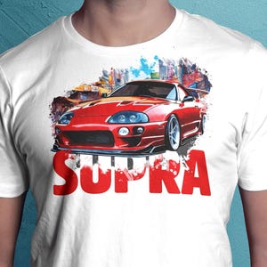 以下が含まれることがあります： 赤いSupraスポーツカーのデザインと街並みを背景にした白いTシャツ。「SUPRA」の文字が車の真下に赤色でプリントされています。柔らかい素材で作られたシャツです。