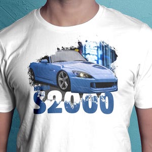 May include: White t-shirt featuring a blue Honda S2000 sports car graphic. The car is depicted with a city backdrop and the text "HONDA $2000" in blue. The shirt is a classic crew neck.