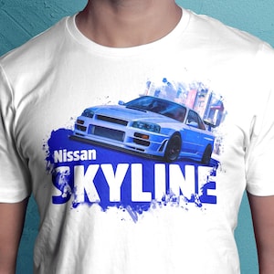 May include: White t-shirt featuring a blue Nissan Skyline car graphic. The design includes the word "Nissan" above the word "SKYLINE" in large, white, bold letters on a blue background. The car is light blue.