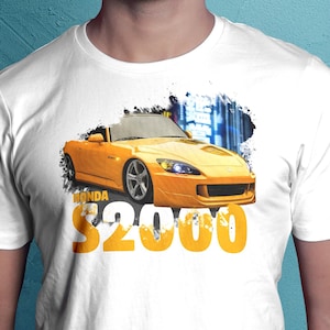 May include: White t-shirt featuring a yellow Honda S2000 sports car graphic. The car is set against a blurred cityscape background, with the word "HONDA" and the number "$2000" in large orange letters.