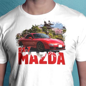 Könnte beinhalten: Weißes T-Shirt mit einer roten Mazda RX-7 Sportwagen-Grafik. Das Design zeigt das Auto, japanische Architektur und Kirschblüten. Die Texte "RX-7" und "MAZDA" sind rot.