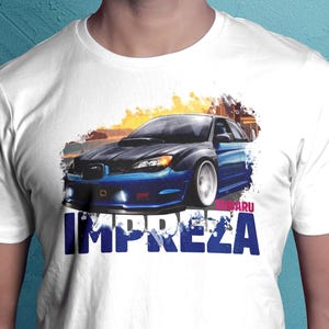 May include: White t-shirt featuring a blue Subaru Impreza car graphic. The design includes the word "IMPREZA" in large blue letters and the Subaru logo. The car is depicted with a sunset background.