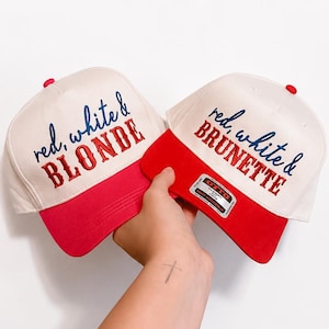 May include: Two baseball caps with off-white crowns and red brims. The left cap features embroidered text reading red, white & BLONDE in blue and red. The right cap features embroidered text reading red, white & BRUNETTE in blue and red.