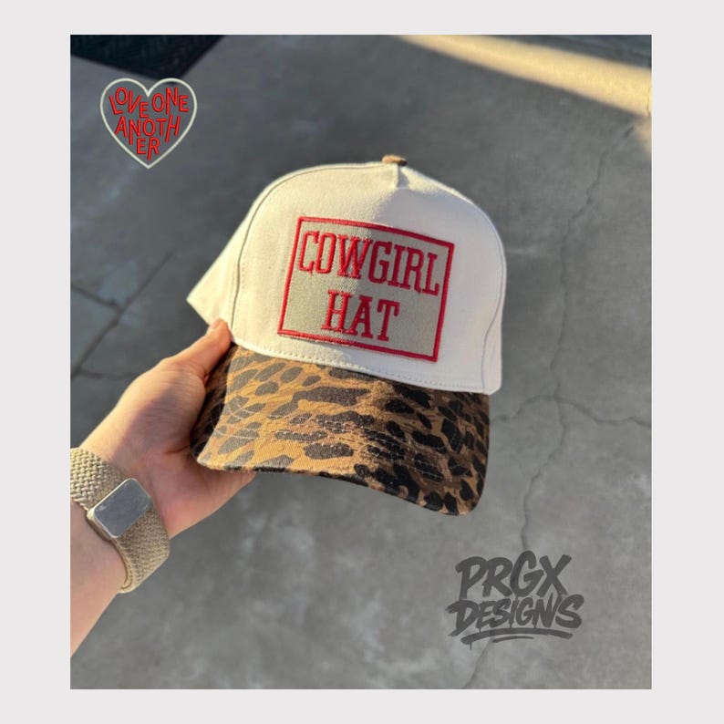 May include: A baseball cap with a cream-colored crown and a brown and black leopard print brim. The front features a rectangular patch with a red border and the red embroidered text COWGIRL HAT. A heart-shaped logo with red text is visible.