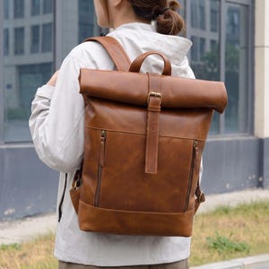 May include: A brown leather roll-top backpack featuring a vertical strap with a metal buckle closure, a top handle, and two front vertical zipped pockets with matching leather pull tabs. The bag has adjustable shoulder straps and a structured base.