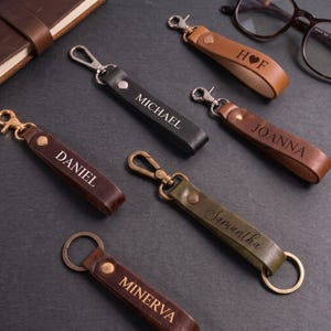May include: A collection of six leather keychains in black, brown, and olive green, each featuring a metal clasp or ring, a rivet, and personalized engravings including the names Michael, Daniel, Joanna, Samantha, Minerva, and H heart F.