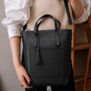 Op de afbeelding: Een zwarte leren tas met twee handvatten, een afneembare schouderriem met goudkleurige metalen hardware en een groot rechthoekig voorvak. Een decoratieve leren kwast hangt aan een van de bovenste handvatten van de tas.