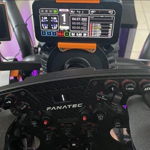May include: A black Fanatec racing steering wheel with carbon fiber texture, multiple buttons, and rotary switches. A digital display screen is mounted above the wheel, showing telemetry data, lap times, and race rankings with white, red, and orange text.