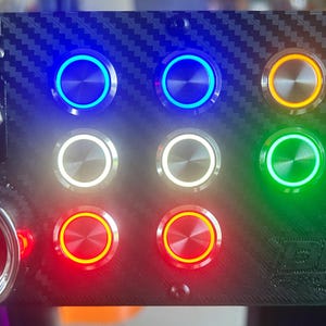 Puede incluir: Una caja de botones de fibra de carbono negra con un botón de arranque rojo grande, un interruptor de palanca rojo y ocho botones pulsadores iluminados en azul, blanco, verde, naranja y rojo. El dispositivo incluye un logotipo DLA en relieve.