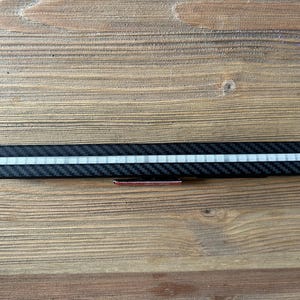 May include: A long, rectangular, black carbon fiber-textured accessory featuring a central horizontal strip of small, white, square-shaped segments. A small black mounting bracket with a red adhesive backing is attached to the center underside.