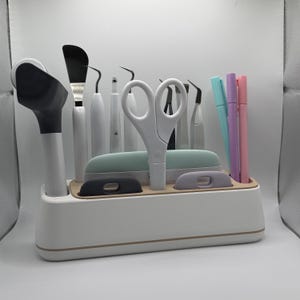 Peut inclure: Un organisateur de bureau rectangulaire blanc avec une base intérieure beige contenant divers outils de loisirs créatifs, dont une paire de ciseaux blancs, un cutter rotatif noir et blanc, une spatule, des pics en métal, des pinces et trois stylos.