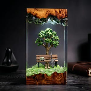 Könnte beinhalten: Eine rechteckige Harz- und Holzlampe mit einer Miniaturszene, bestehend aus einem grünen Baum, einer Holzbank, einem Helm, einem Gewehr und einem Paar Stiefel auf einem grasbewachsenen Untergrund. Ober- und Unterteil bestehen aus Naturholz mit Maserung.