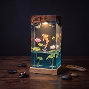 Peut inclure: Une lampe rectangulaire en résine avec une base et un sommet en bois naturel, renfermant une scène d'étang miniature avec un poisson koi blanc et orange, des fleurs de lotus roses et des nénuphars verts, éclairée par une lumière chaude.