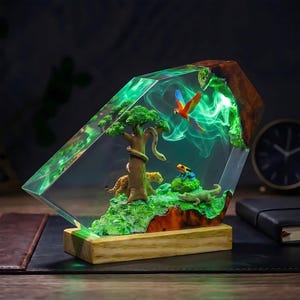 May include: A decorative resin lamp featuring a jungle scene with a leopard, a snake on a tree, a frog, a crocodile, and a flying macaw. The clear resin block has green internal lighting and is mounted on a rectangular light-colored wooden base.