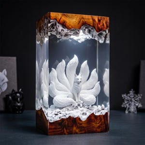 Peut inclure: Une lampe rectangulaire avec une figurine de renard blanc à neuf queues encapsulée dans de la résine transparente entre deux morceaux de bois brun foncé poli. Le renard repose sur une base blanche texturée et le bois supérieur est irrégulier.