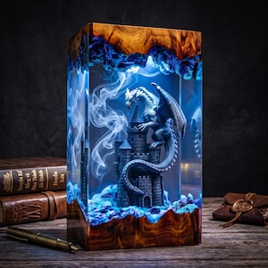 Puede incluir: Una lámpara rectangular hecha de madera natural y resina azul translúcida. Dentro de la resina, un dragón gris se posa sobre una torre de castillo de piedra. El interior está iluminado con una luz azul brillante, creando un efecto de humo.