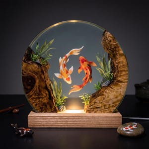 Può includere: Una lampada circolare in resina con due pesci koi arancioni, bianchi e neri, sospesi all'interno di una cornice trasparente affiancata da pezzi di legno naturale e piante acquatiche verdi. La lampada poggia su una base rettangolare in legno.