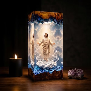 May include: A rectangular resin and wood lamp featuring a central figure of Jesus with arms outstretched, standing on a cloud. The interior displays a blue sky with white clouds, encased in clear resin with dark wood top and bottom borders.