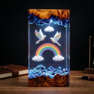 Puede incluir: Un bloque de resina rectangular con vetas de madera natural en la parte superior e inferior. Dentro de la resina transparente, dos palomas blancas con alas de arcoíris vuelan sobre un arcoíris, flanqueadas por nubes blancas y texturas azules.