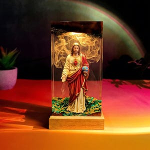 May include: A rectangular resin lamp featuring a figurine of Jesus Christ wearing a red and white robe, holding a blue orb, and standing on a green textured base. The interior contains swirling golden light effects and a dark wooden top.