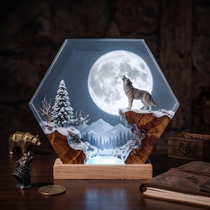 May include: A hexagonal resin and wood lamp featuring a backlit full moon, a miniature snow-covered pine tree, and a wolf figurine howling on a rocky cliff. The scene includes a mountain landscape silhouette and a light-colored wooden base stand.