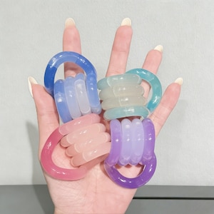 May include: A hand displaying five colorful, translucent rings. Each ring features a large, curved band connected to a stack of smaller, rounded bands. The rings are in shades of blue, pink, purple, and green.