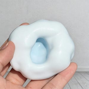 May include: A light blue slime with a white outer layer, held in a hand. The slime has a cloud-like appearance with a hole in the center revealing a lighter blue color. The texture appears smooth and soft.