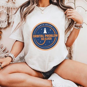 May include: White t-shirt with a circular graphic featuring a rocket ship and the text "EARTH-BASED ORBITAL PROBLEM SOLVER" in blue and orange. The design is centered on the front of the shirt.