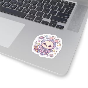 May include: A sticker depicting a cartoon bunny astronaut with a rocket, surrounded by stars and planets. The sticker has a white border and is placed on a laptop. The bunny is wearing a pink and purple spacesuit.