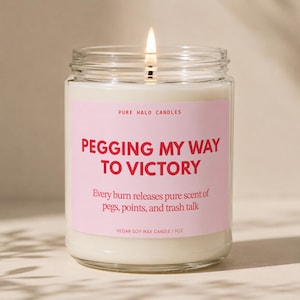 May include: A clear glass jar candle with a lit wick and a light pink label. The label reads PURE HALO CANDLES, PEGGING MY WAY TO VICTORY, Every burn releases pure scent of pegs, points, and trash talk, and VEGAN SOY WAX CANDLE | 255g in red and black text.