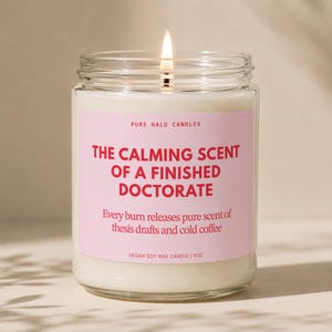 May include: A lit soy wax candle in a clear glass jar with a pink label. The text reads: PURE HALO CANDLES, THE CALMING SCENT OF A FINISHED DOCTORATE, Every burn releases pure scent of thesis drafts and cold coffee, VEGAN SOY WAX CANDLE | 9OZ.