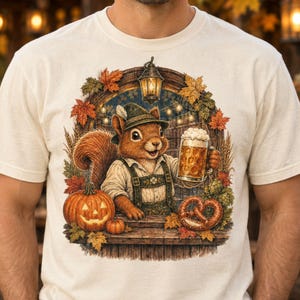 May include: A cream-colored t-shirt featuring a graphic of a squirrel wearing a traditional alpine hat and suspenders, holding a large beer mug. The scene includes a jack-o'-lantern, a pretzel, autumn leaves, and a hanging lantern.