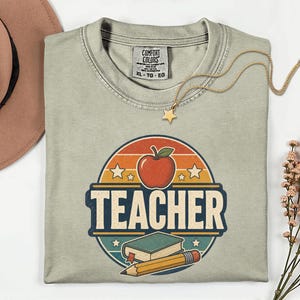 Folded Sandstone teacher shirt featuring a retro TEACHER graphic with apple, book, pencil, stars and vintage distressed school design.