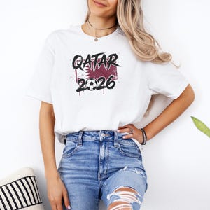 White t-shirt mockup featuring a bold Qatar 2026 soccer graphic with maroon flag-inspired design, grunge lettering, and soccer ball detail.