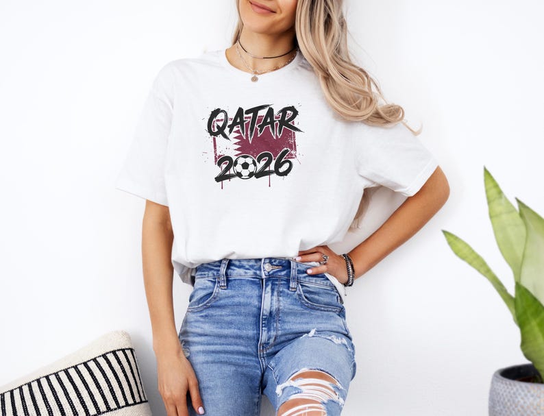 White t-shirt mockup featuring a bold Qatar 2026 soccer graphic with maroon flag-inspired design, grunge lettering, and soccer ball detail.