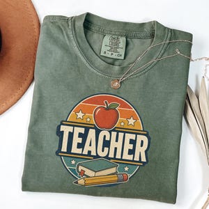 Folded Moss teacher shirt featuring a vintage TEACHER design with apple, book, pencil, stars and retro classroom colors.