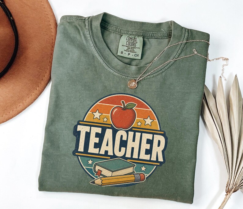 Folded Moss teacher shirt featuring a vintage TEACHER design with apple, book, pencil, stars and retro classroom colors.