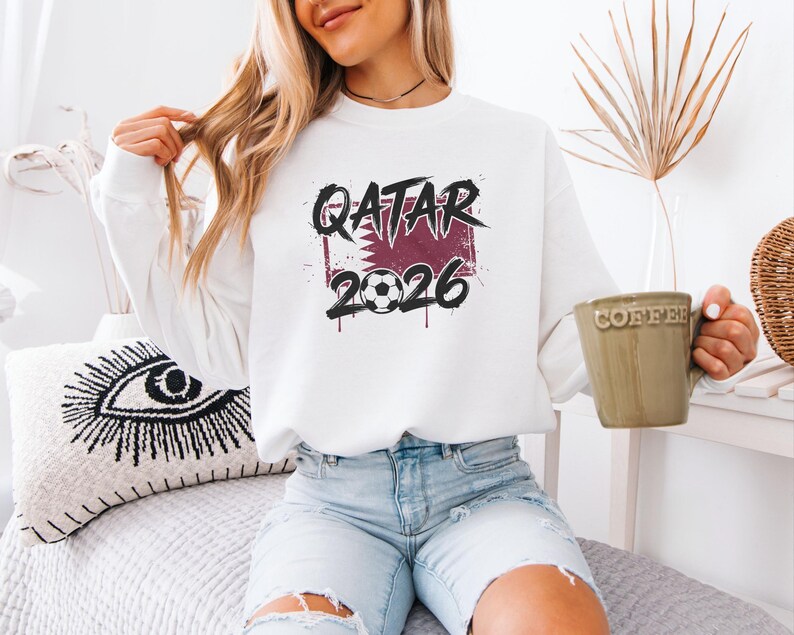White sweatshirt mockup featuring a Qatar 2026 soccer graphic with maroon flag-inspired background, bold grunge text, and soccer ball detail.