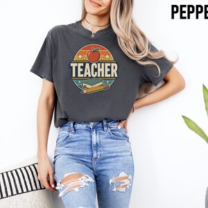 Woman wearing a Pepper teacher shirt with bold retro TEACHER lettering, apple, book and pencil graphic on the front.