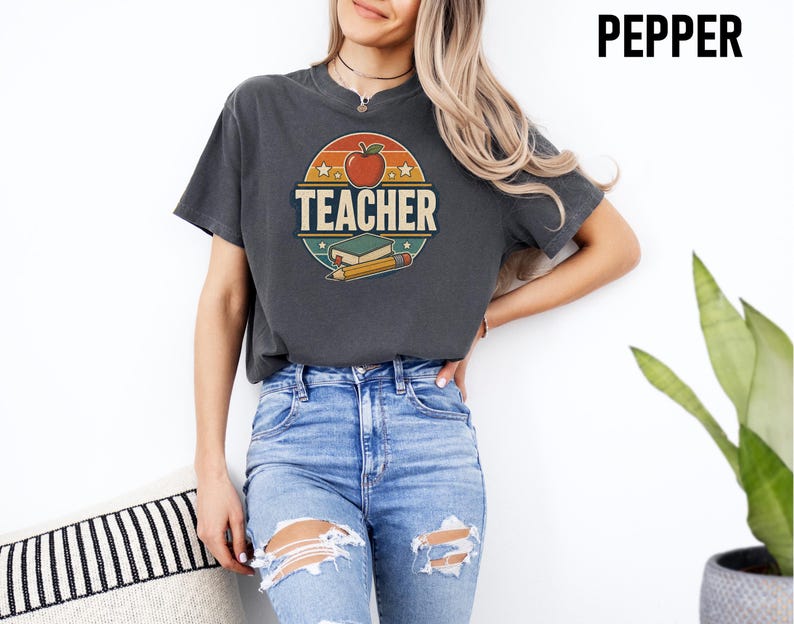 Woman wearing a Pepper teacher shirt with bold retro TEACHER lettering, apple, book and pencil graphic on the front.