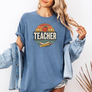 Woman wearing a Blue Jean teacher shirt with vintage TEACHER lettering, apple, book, pencil and distressed classroom graphic.