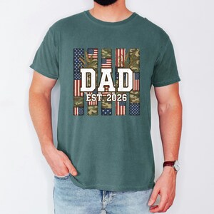 Puede incluir: Una camiseta de manga corta color verde azulado oscuro con un estampado gráfico que incluye el texto DAD EST. 2026 en letras blancas. El diseño presenta franjas verticales con patrones de bandera estadounidense y camuflaje tras el texto.