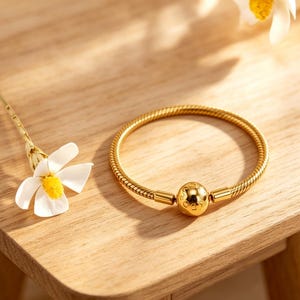 Puede incluir: Una pulsera de cadena de serpiente en tono dorado con una cuenta esférica central con texto grabado. La pulsera está colocada junto a una flor blanca con centro amarillo, mostrando un acabado metálico liso y un diseño circular flexible.