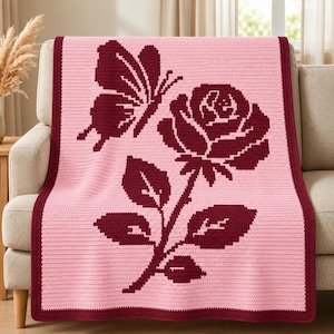May include: A rectangular crochet blanket featuring a light pink background with a dark maroon border. The center displays a large, dark maroon graphic of a blooming rose with a stem and leaves, accompanied by a butterfly positioned above.