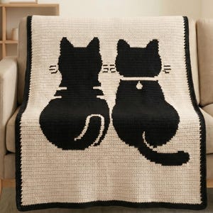 Könnte beinhalten: Eine cremefarbene Häkeldecke, die über einem Sofa drapiert ist. Die Decke zeigt zwei große schwarze Katzensilhouetten, eine davon mit Halsband. Die Decke hat einen schwarzen Rand und besteht aus einem weichen, strukturierten Material.