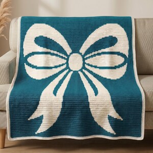 May include: A rectangular crochet blanket featuring a large, centered cream-colored bow graphic against a solid teal background. The blanket has a simple cream-colored border that frames the entire piece, showcasing a consistent knit texture.